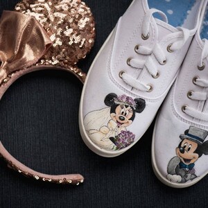 May include: A pair of white canvas shoes with hand-painted images of Mickey and Minnie Mouse dressed in wedding attire. The shoes are tied with white laces and have a blue and white polka dot lining. A rose gold headband with a bow and sequins is also pictured.