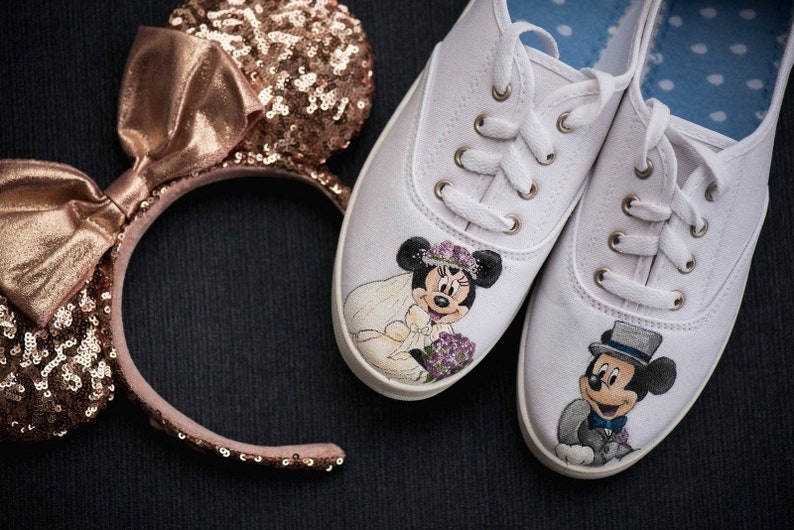 May include: A pair of white canvas shoes with hand-painted images of Mickey and Minnie Mouse dressed in wedding attire. The shoes are tied with white laces and have a blue and white polka dot lining. A rose gold headband with a bow and sequins is also pictured.