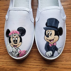 May include: A pair of white canvas shoes with hand-painted images of Mickey Mouse and Minnie Mouse dressed in wedding attire. Mickey is wearing a black tuxedo with a top hat and bow tie. Minnie is wearing a white dress with a pink flower crown and veil.