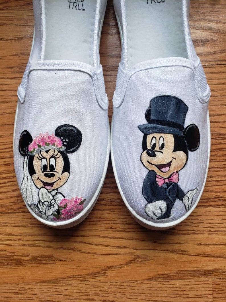 May include: A pair of white canvas shoes with hand-painted images of Mickey Mouse and Minnie Mouse dressed in wedding attire. Mickey is wearing a black tuxedo with a top hat and bow tie. Minnie is wearing a white dress with a pink flower crown and veil.