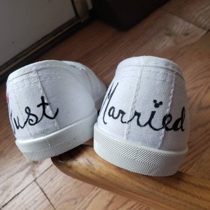 May include: A pair of white canvas shoes with black embroidery. One shoe says "Just" and the other says "Married".