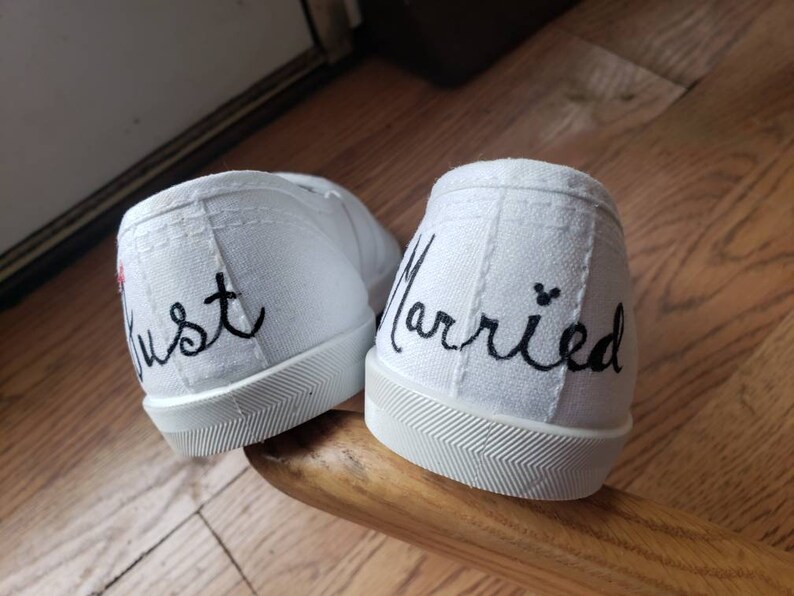 May include: A pair of white canvas shoes with black embroidery. One shoe says "Just" and the other says "Married".