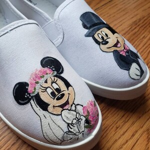 May include: A pair of white canvas slip-on shoes with hand-painted images of Mickey and Minnie Mouse dressed in wedding attire. Minnie Mouse is wearing a white dress with a veil and a floral crown. Mickey Mouse is wearing a tuxedo and a top hat.
