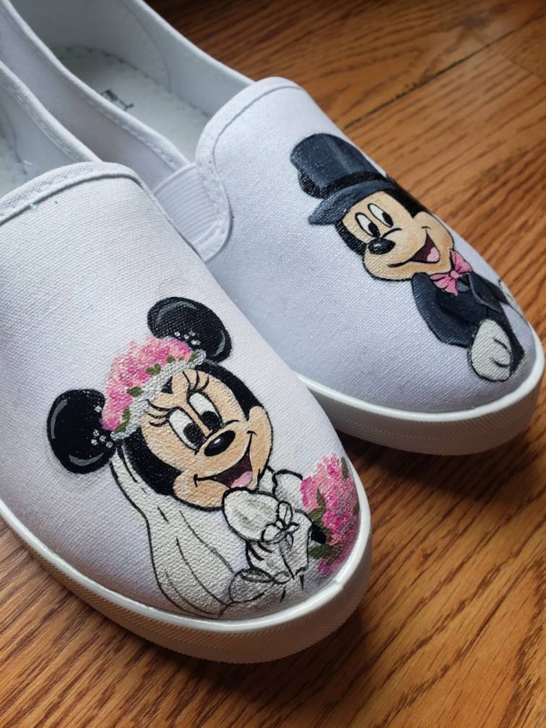May include: A pair of white canvas slip-on shoes with hand-painted images of Mickey and Minnie Mouse dressed in wedding attire. Minnie Mouse is wearing a white dress with a veil and a floral crown. Mickey Mouse is wearing a tuxedo and a top hat.