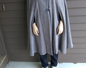 May include: A gray tweed cape with a rounded collar and decorative toggle closures. The cape is knee-length and has slits for the arms. The person is wearing black pants and white sneakers with black stripes.