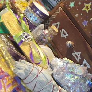 May include: A collection of crystals, a wooden wand, a sage smudge stick, and a small glass bottle with a cork stopper. The items are arranged on a purple and green fabric with a wooden box in the background. The box has a drawer with a white star design and the text "Shine Bright".