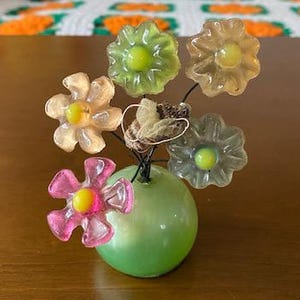 May include: A decorative floral arrangement featuring six translucent, colorful flowers with yellow centers. The flowers are arranged in a green, spherical vase. The arrangement sits on a wooden surface.