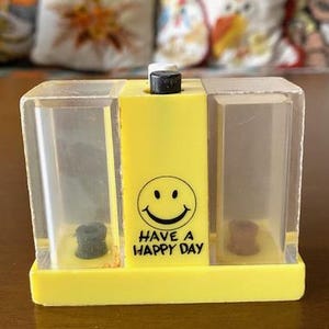 May include: A yellow and clear plastic container with a smiley face and the text "Have a Happy Day." The container has two compartments with small, dark objects inside. A black and white button is on top.