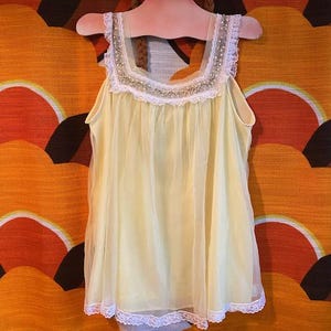 May include: A vintage yellow nightgown with white lace trim, displayed on a wooden doll-shaped hanger. The nightgown has a square neckline and a sheer overlay. The hanger is painted with a doll face, featuring blue eyes and braided brown hair.