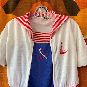 May include: White and red striped sailor shirt with a blue and white striped top underneath. The shirt has a zipper closure and a hood. The shirt has a red and white striped trim around the bottom and sleeves. The shirt has a small sailboat embroidered on the right side and an anchor embroidered on the left side.