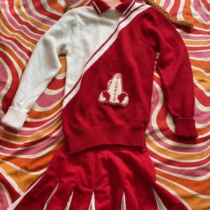 May include: A vintage cheerleader outfit in red and white, featuring a sweater with a "Michelle" patch and a pleated skirt. The outfit is displayed with a decorative doll head with pigtails, set against a colorful, retro-style background.