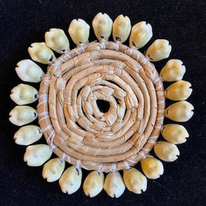 May include: A circular decorative piece featuring a woven, spiral-shaped center made of natural fibers. Cream-colored seashells are evenly spaced around the perimeter, creating a sunburst effect. The shells have a glossy finish.