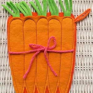 May include: A felt carrot-shaped pouch with five orange carrots, green tops, and a pink string tied around the middle. The pouch has a loop on the side. The design is reminiscent of Easter or spring.