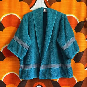 May include: Teal terry cloth robe with a shawl collar and short sleeves. The robe has a white and gray striped detail on the sleeves and hem. The robe is hanging on a white hanger against a retro orange floral background.
