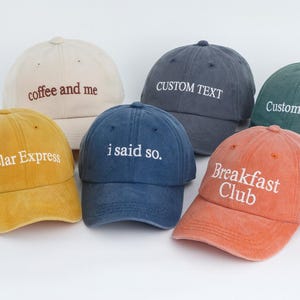 May include: A collection of six baseball caps in various colors, including yellow, beige, navy, and orange. Each cap features embroidered text, such as "coffee and me," "i said so," and "Breakfast Club."