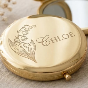 May include: A gold-colored compact mirror with the name "Chloe" engraved on the lid, along with a floral design. The mirror is open, revealing a reflective surface. A second, similar compact is visible in the background.