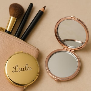 May include: A gold compact mirror with the name "Laila" engraved on the front. The mirror is open and shows a reflection. The mirror is next to a beige zippered pouch with three makeup brushes inside.