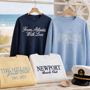 May include: Four crewneck sweatshirts in navy, light blue, yellow, and white, featuring embroidered text including "From Atlantis With Love," "Slow Morning Club EST. 2025," "THE MILLERS Palm Circle EST. 2025," and "NEWPORT Beach Club," alongside a white captain's hat.