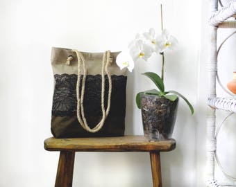 May include: A beige canvas tote bag with black lace trim and rope handles. The bag is sitting on a wooden stool. A white orchid plant is in a clear pot behind the bag.