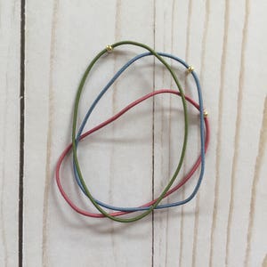 Könnte beinhalten: Drei elastische Haargummis mit goldenen Perlen. Die Haargummis sind grün, blau und rosa.