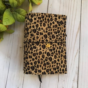 Puede incluir: Una cubierta de cuaderno de tela con estampado de leopardo marrón y negro con un dije de corazón dorado.