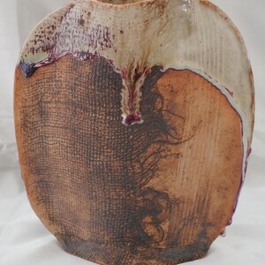 May include: A flattened ceramic vase with a textured brown surface featuring a cross-hatched pattern on one side and a smooth, cream-colored glaze dripping down from the top rim. The base is rounded and the body has a distinct, organic shape.