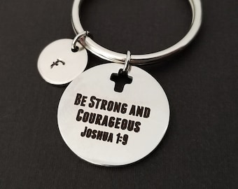 May include: Silver keychain with a large round pendant engraved with the text "BE STRONG AND COURAGEOUS JOSHUA 1:9". Includes a smaller round charm with the letter "f" and a silver key ring. The background is black.