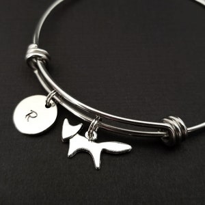 May include: Silver-toned adjustable bangle bracelet with a fox charm and a round charm with the letter "R". The bracelet is on a black surface. The fox charm is a stylized outline.