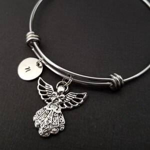 May include: A silver-colored bangle bracelet with an angel charm and a small round charm with the letter "N". The angel charm has detailed wings and a flowing gown. The bracelet is on a dark background.