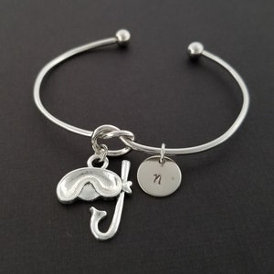 May include: Silver bangle bracelet with a charm of a snorkel and swim mask and a round charm with the letter "N" engraved on it.