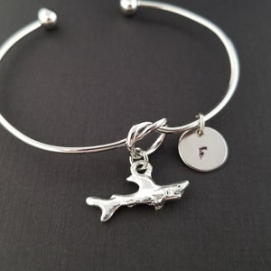 May include: Silver bangle bracelet with a knotted charm and a shark charm. The bracelet also has a small round charm with the letter "F" engraved on it.