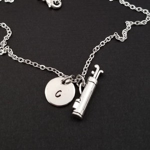 May include: A silver necklace with a golf bag charm and a round charm with the letter "C" engraved on it.