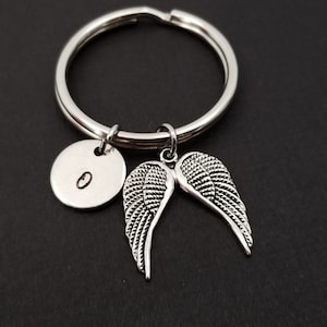 May include: Silver-toned keychain featuring a round key ring, a circular charm with the letter "O", and a pair of detailed angel wings. The wings are intricately designed with feather detailing. The keychain is likely made of metal.
