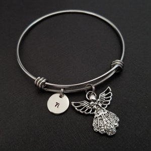 May include: A silver-colored bangle bracelet with two charms. One charm is a small, round disc with the letter "n" engraved on it. The other charm is a detailed angel pendant with outstretched wings. The bracelet is on a black surface.