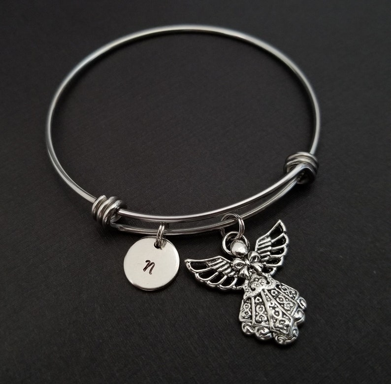May include: A silver-colored bangle bracelet with two charms. One charm is a small, round disc with the letter "n" engraved on it. The other charm is a detailed angel pendant with outstretched wings. The bracelet is on a black surface.