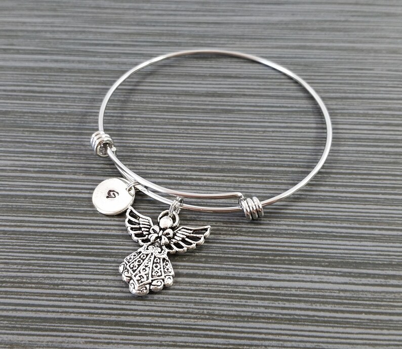 May include: A silver-toned adjustable bangle bracelet with an angel charm and a small round charm with the letter "S". The angel charm has detailed wings and a floral-like design on the dress.
