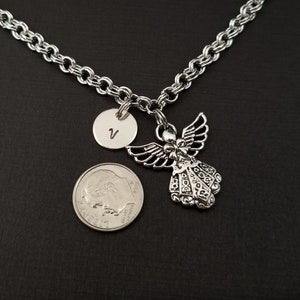 Peut inclure: Un collier en cha&icirc;ne argent&eacute;e avec un pendentif ange en argent et un pendentif rond en argent grav&eacute; de la lettre "V". Le collier est pr&eacute;sent&eacute; sur une surface noire.