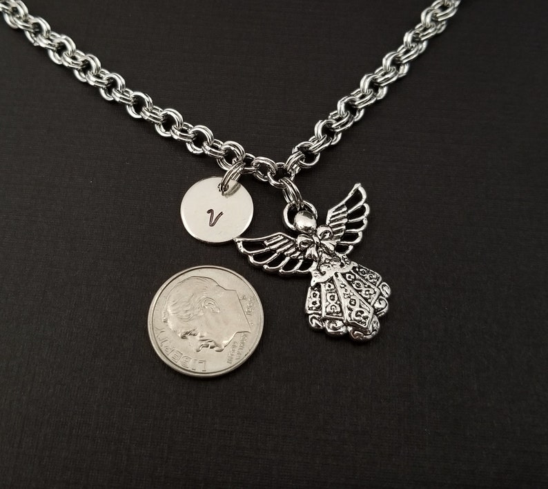 Peut inclure: Un collier en cha&icirc;ne argent&eacute;e avec un pendentif ange en argent et un pendentif rond en argent grav&eacute; de la lettre "V". Le collier est pr&eacute;sent&eacute; sur une surface noire.