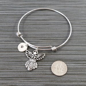 May include: A silver-colored bangle bracelet with an angel charm and a small round charm with the letter "G". The angel charm has detailed wings and a decorative dress. A U.S. dime is next to the bracelet for size comparison.