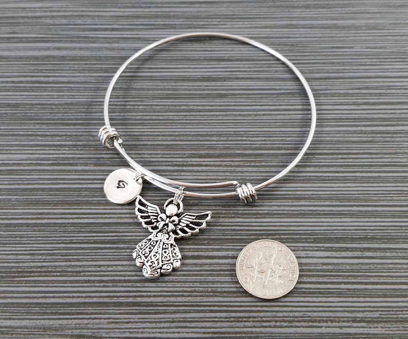 May include: A silver-colored bangle bracelet with an angel charm and a small round charm with the letter "G". The angel charm has detailed wings and a decorative dress. A U.S. dime is next to the bracelet for size comparison.