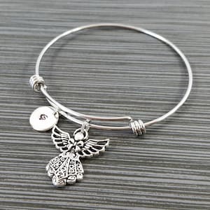 May include: Silver-toned expandable bangle bracelet featuring a detailed angel charm and a small round charm with the letter "G". The angel charm has detailed wings and a decorative dress. The bracelet is displayed on a gray surface.