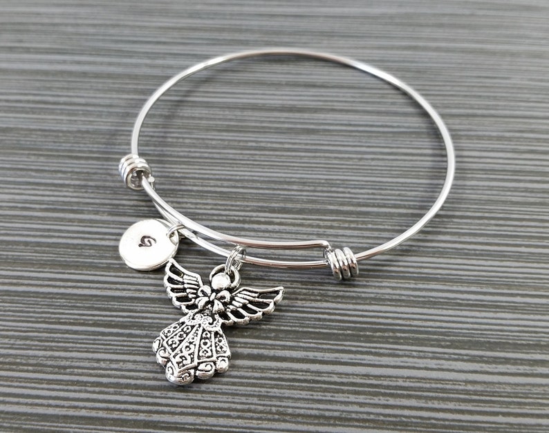 May include: Silver-toned expandable bangle bracelet featuring a detailed angel charm and a small round charm with the letter "G". The angel charm has detailed wings and a decorative dress. The bracelet is displayed on a gray surface.