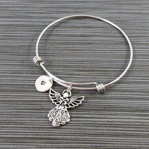 May include: Silver-toned bangle bracelet featuring an angel charm and a small, round charm with a letter. The angel charm has detailed wings and a decorative design. The bracelet is on a gray surface.