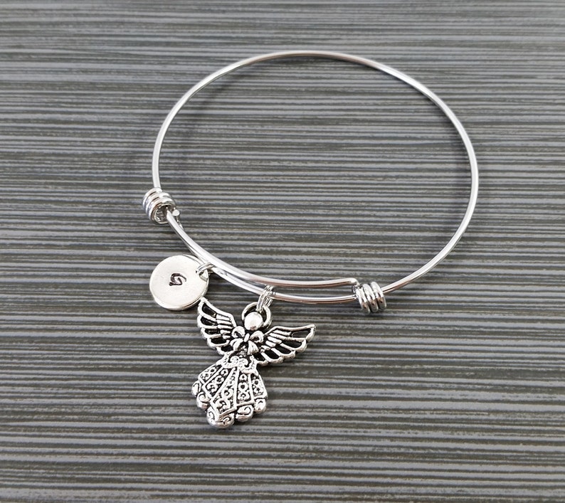 May include: Silver-toned bangle bracelet featuring an angel charm and a small, round charm with a letter. The angel charm has detailed wings and a decorative design. The bracelet is on a gray surface.
