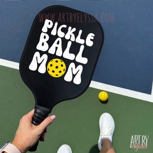 May include: A black pickleball paddle with white text that reads "Pickleball Mom" and a yellow pickleball. The paddle is held by a person's hand on a green and blue pickleball court.