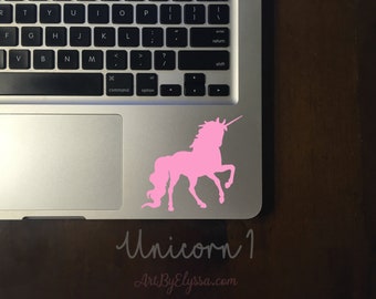 May include: A pink unicorn silhouette sticker on a laptop. The sticker is a solid color and features the mythical creature in a standing pose. The laptop is silver and sits on a dark wooden surface. The text "Unicorn 1" and "ArtByElyssa.com" are also visible.