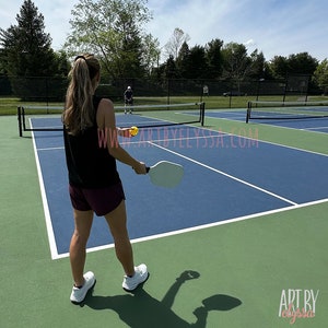 May include: A woman in a black tank top and maroon shorts is playing pickleball on a blue and green court. She is holding a white paddle and is about to hit a yellow ball. A man in a blue shirt is playing against her.