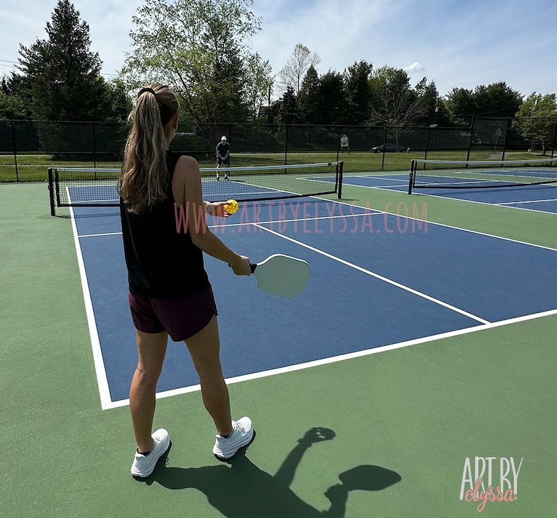 May include: A woman in a black tank top and maroon shorts is playing pickleball on a blue and green court. She is holding a white paddle and is about to hit a yellow ball. A man in a blue shirt is playing against her.
