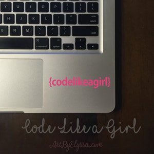 May include: A silver laptop computer with a pink sticker on the trackpad that says "{codelikeagirl}". The laptop is on a wooden surface with the words "code Like a Girl" written in cursive below the laptop.