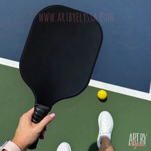May include: A black pickleball paddle with a black grip. The paddle is held by a person on a pickleball court. A yellow pickleball is on the court.